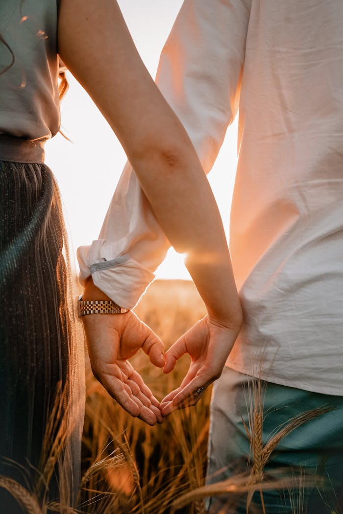 Couple standing in field create heart shape with hands at sunset, symbolizing love.
